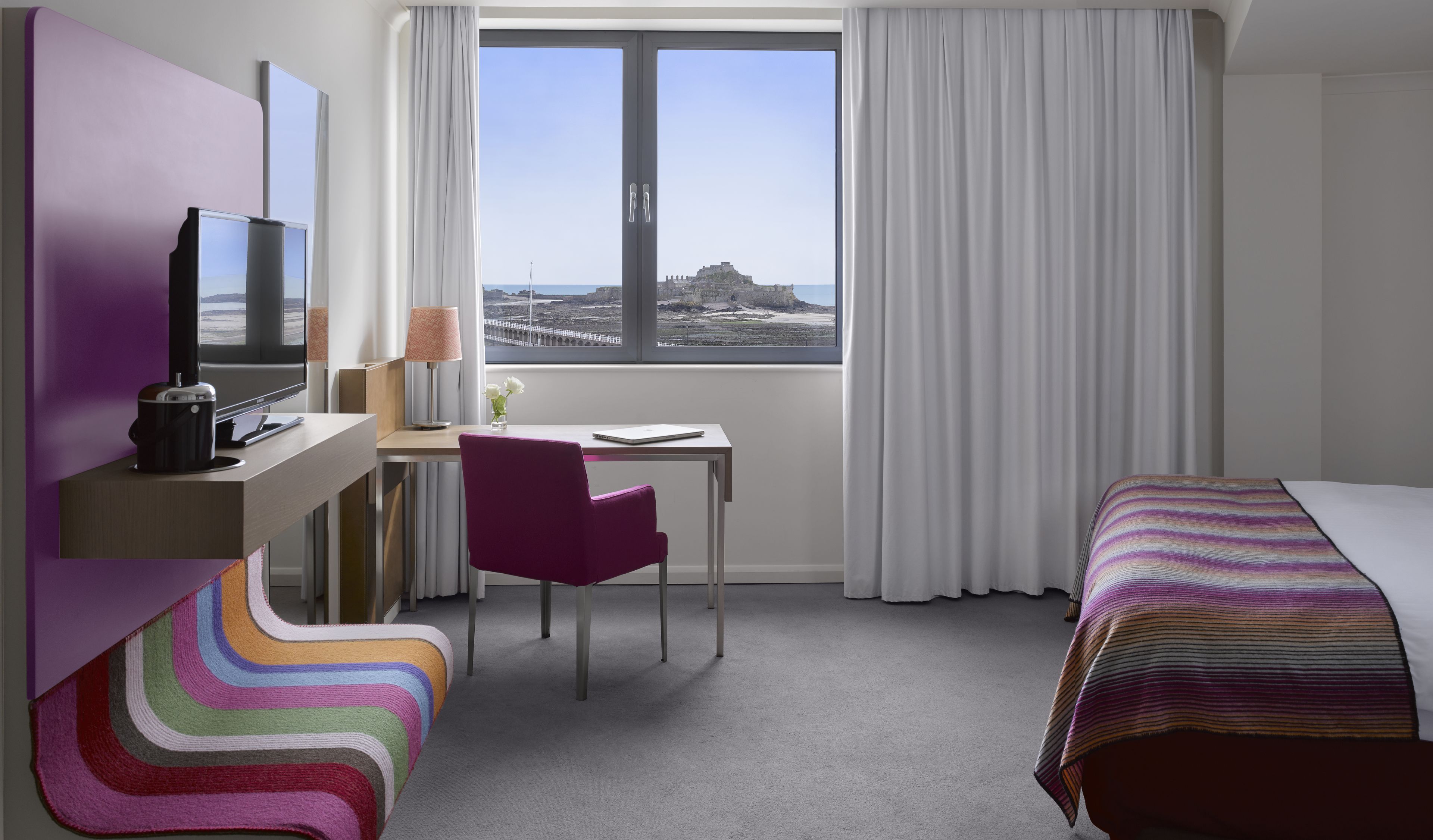 A hotel room with a desk, chair, bed, and a large window offering a view of a coastal landscape.