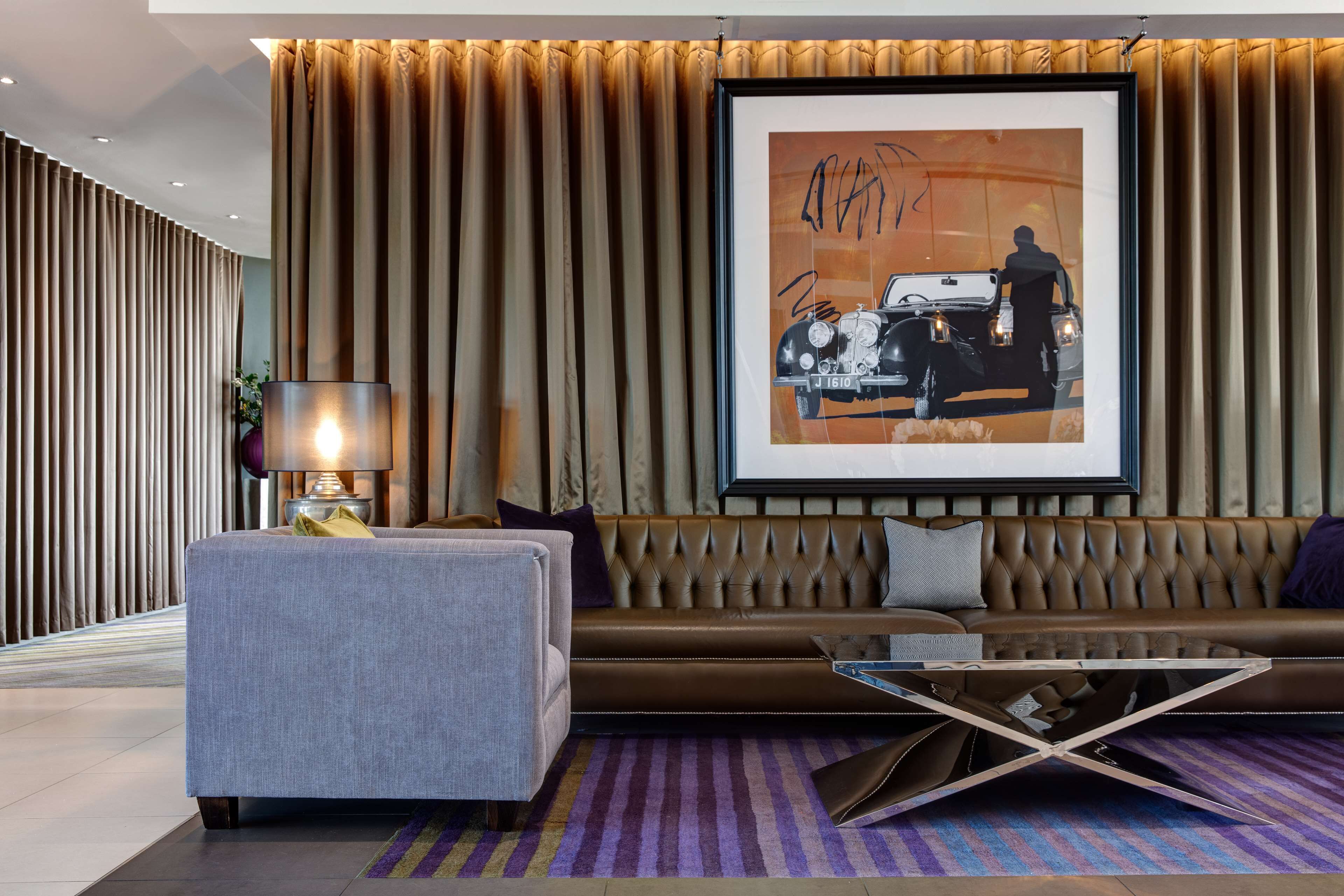 A modern living room with a brown leather sofa, a grey armchair, and a glass coffee table. A framed artwork of a vintage car is displayed on the wall.