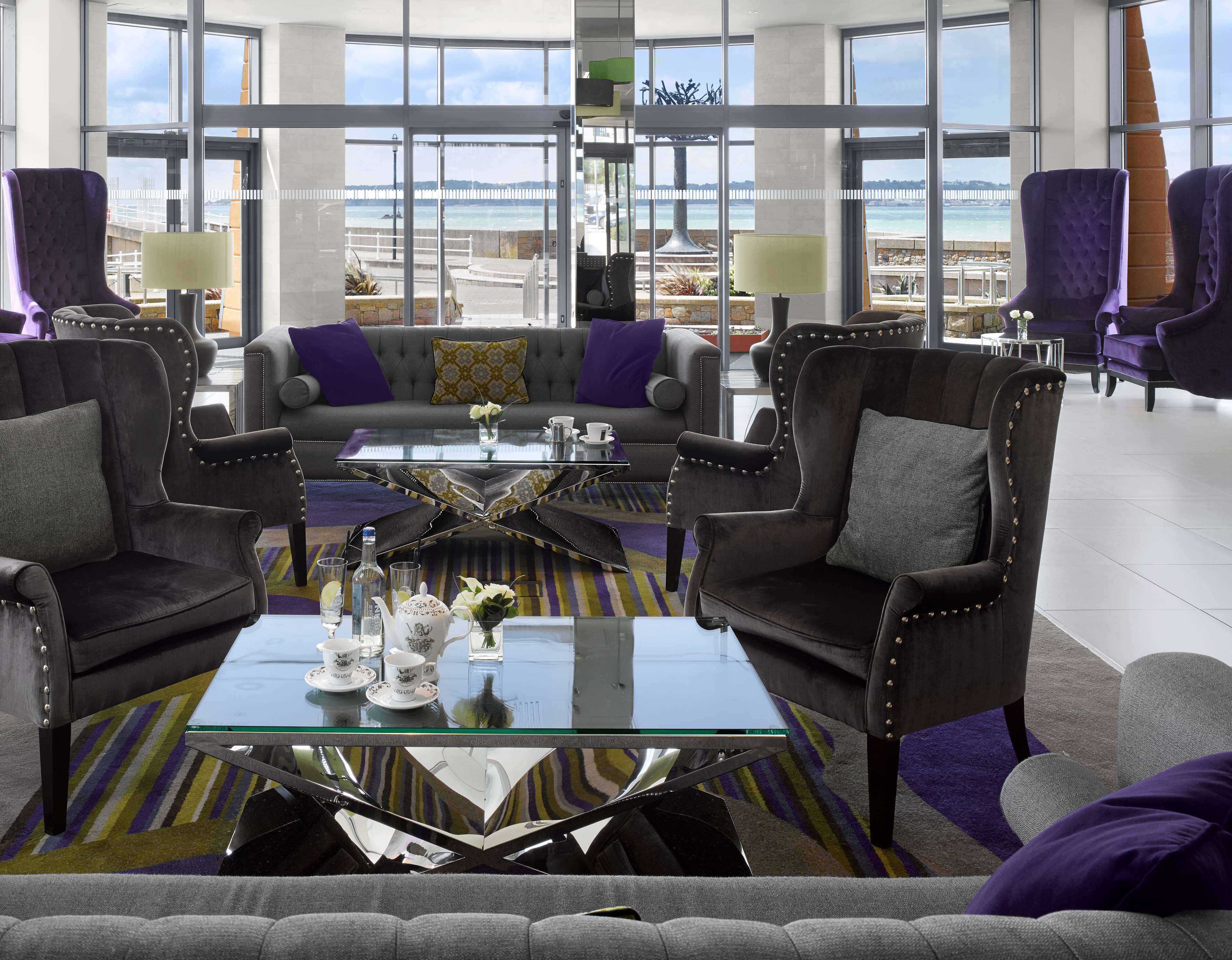 A modern lounge area with a glass coffee table, plush armchairs, and a sofa with a view of the sea.