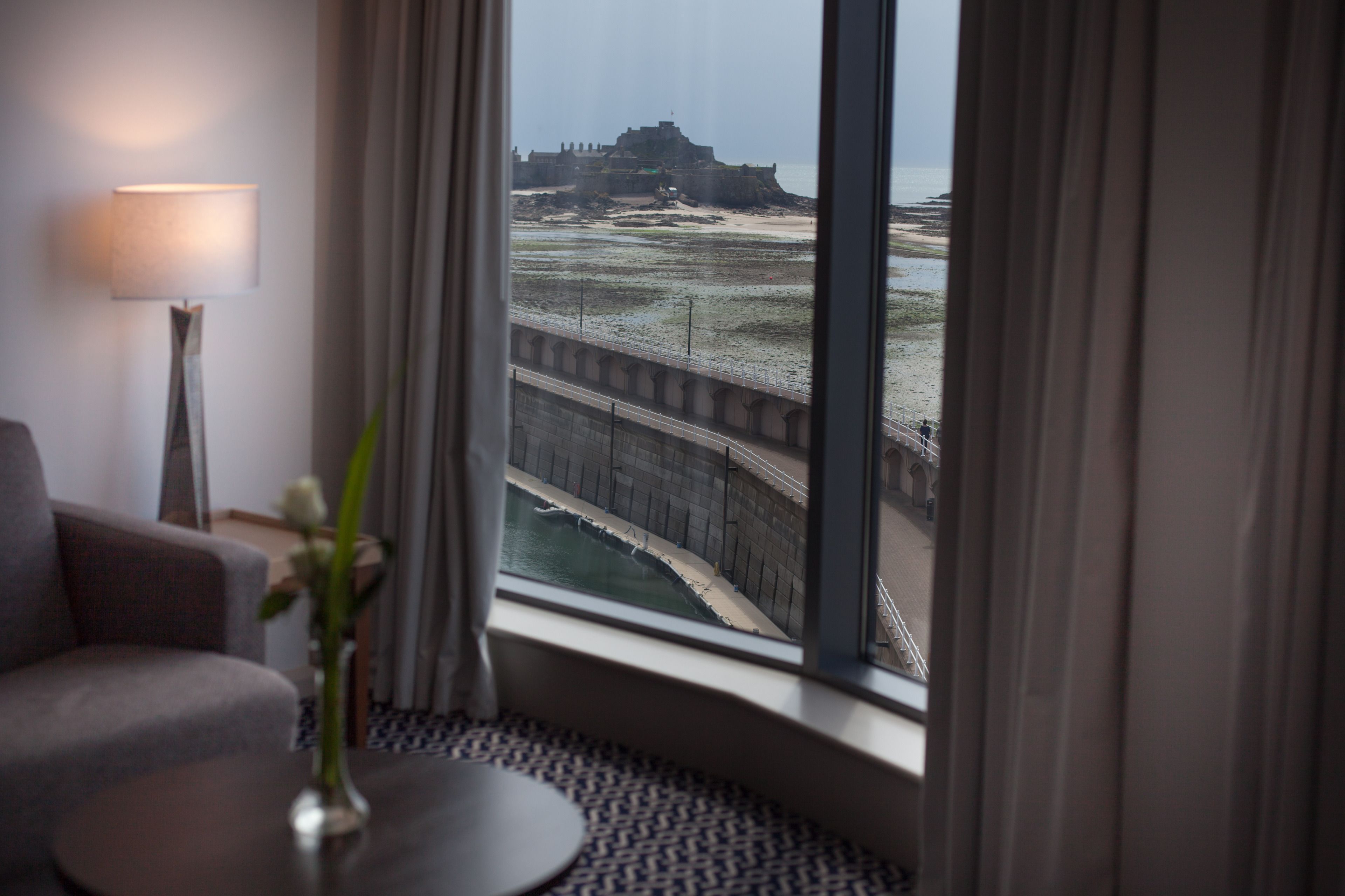 Habitación de hotel con un ventanal grande que ofrece vistas a una fortificación costera y un puente.
