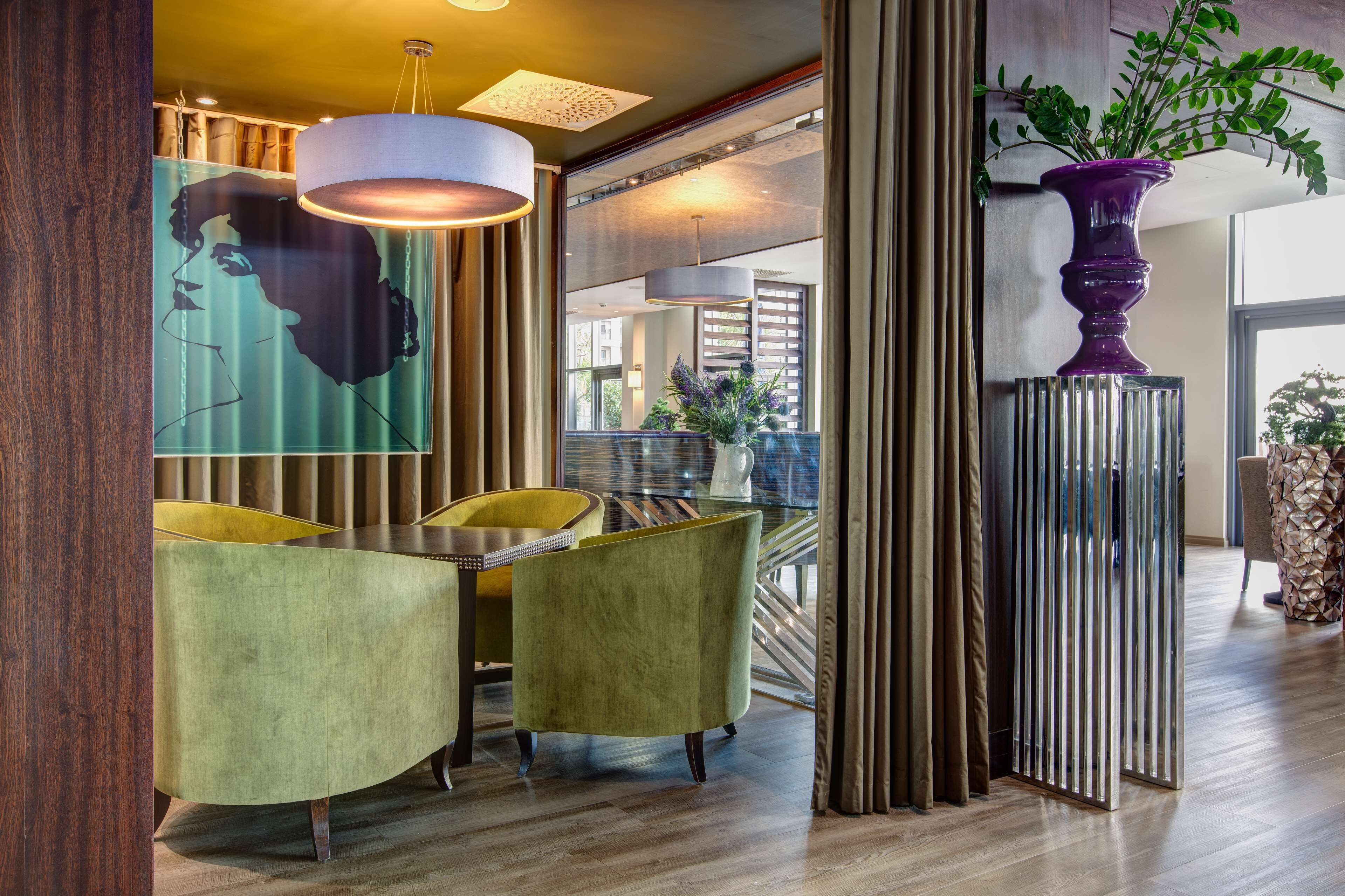 Un interior moderno con un área de comedor que presenta sillas tapizadas de verde y una mesa de cristal, un cuadro abstracto de gran tamaño y un jarrón púrpura con follaje verde.
