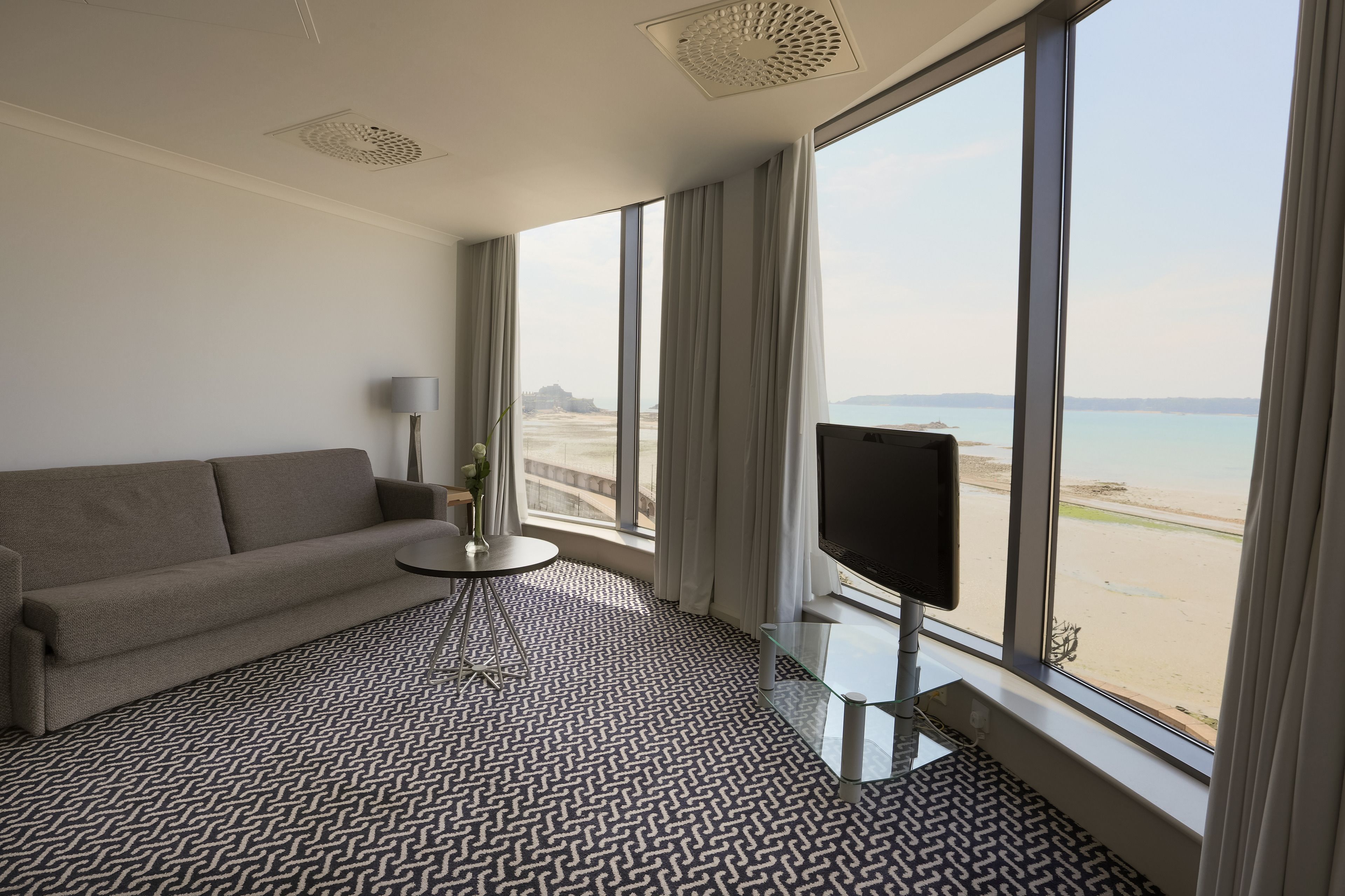 A hotel room with a large window, a sofa, a small table, and a TV.