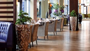 Restaurante com interior moderno, pisos de madeira, cadeiras acolchoadas e diversos vasos de plantas decorativas.