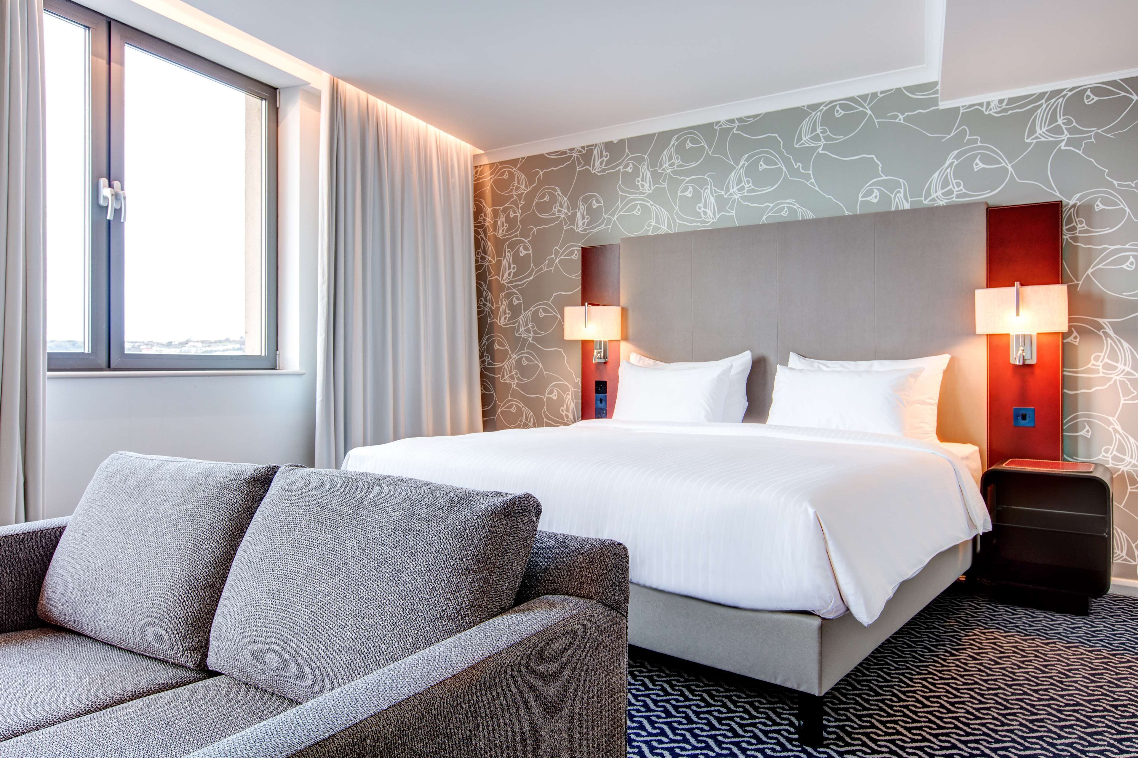 A hotel room with a large bed, two bedside lamps, a sofa, and a patterned wall.