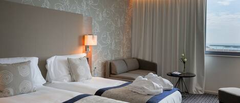 A hotel room with two beds, a sofa, a small table with a vase, and a view of the sea.