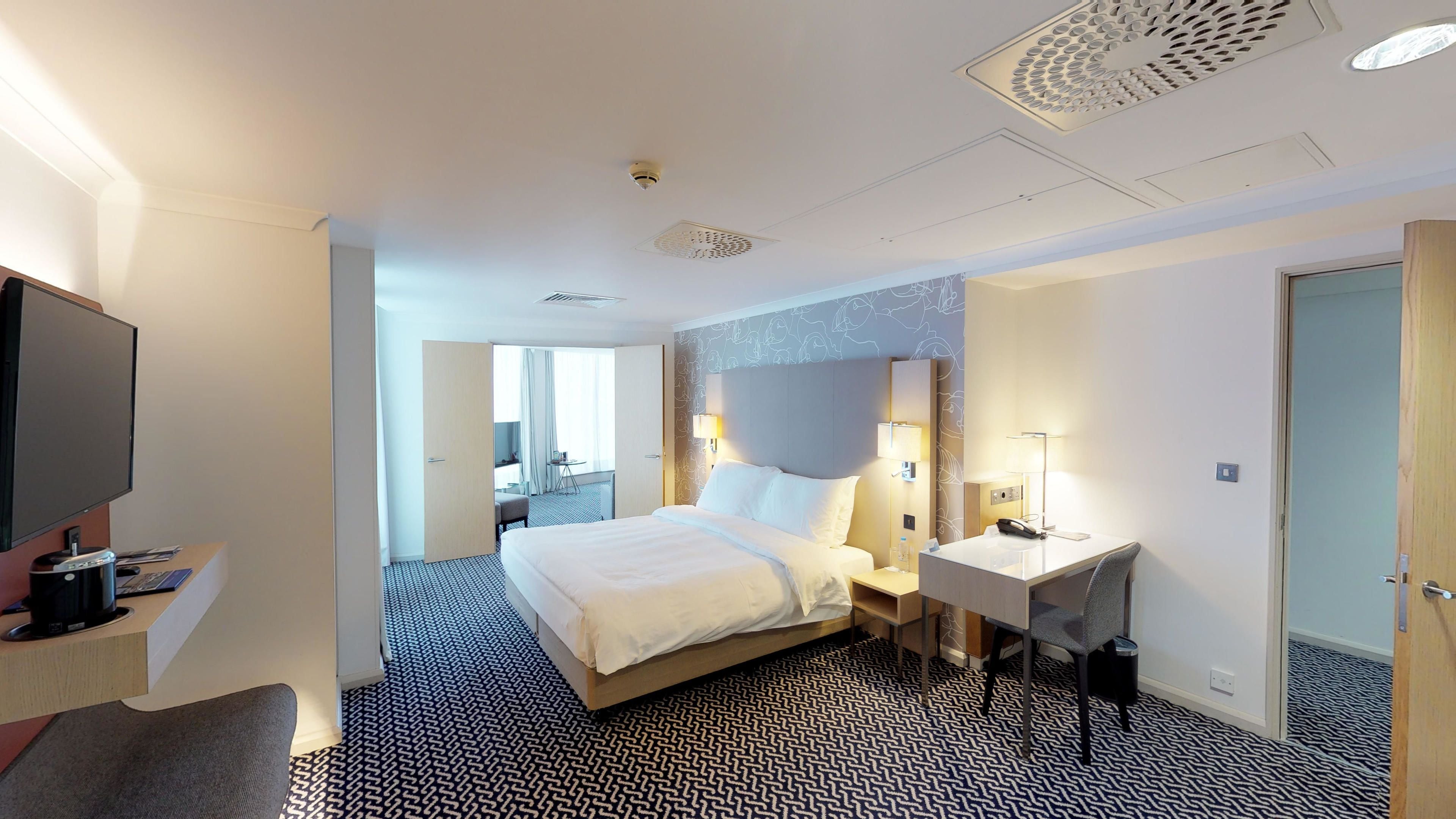 A hotel room with a bed, bedside table, desk, TV, and a bathroom door.