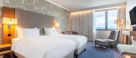 A hotel room with two beds, a large window with curtains, a TV, and a patterned carpet.