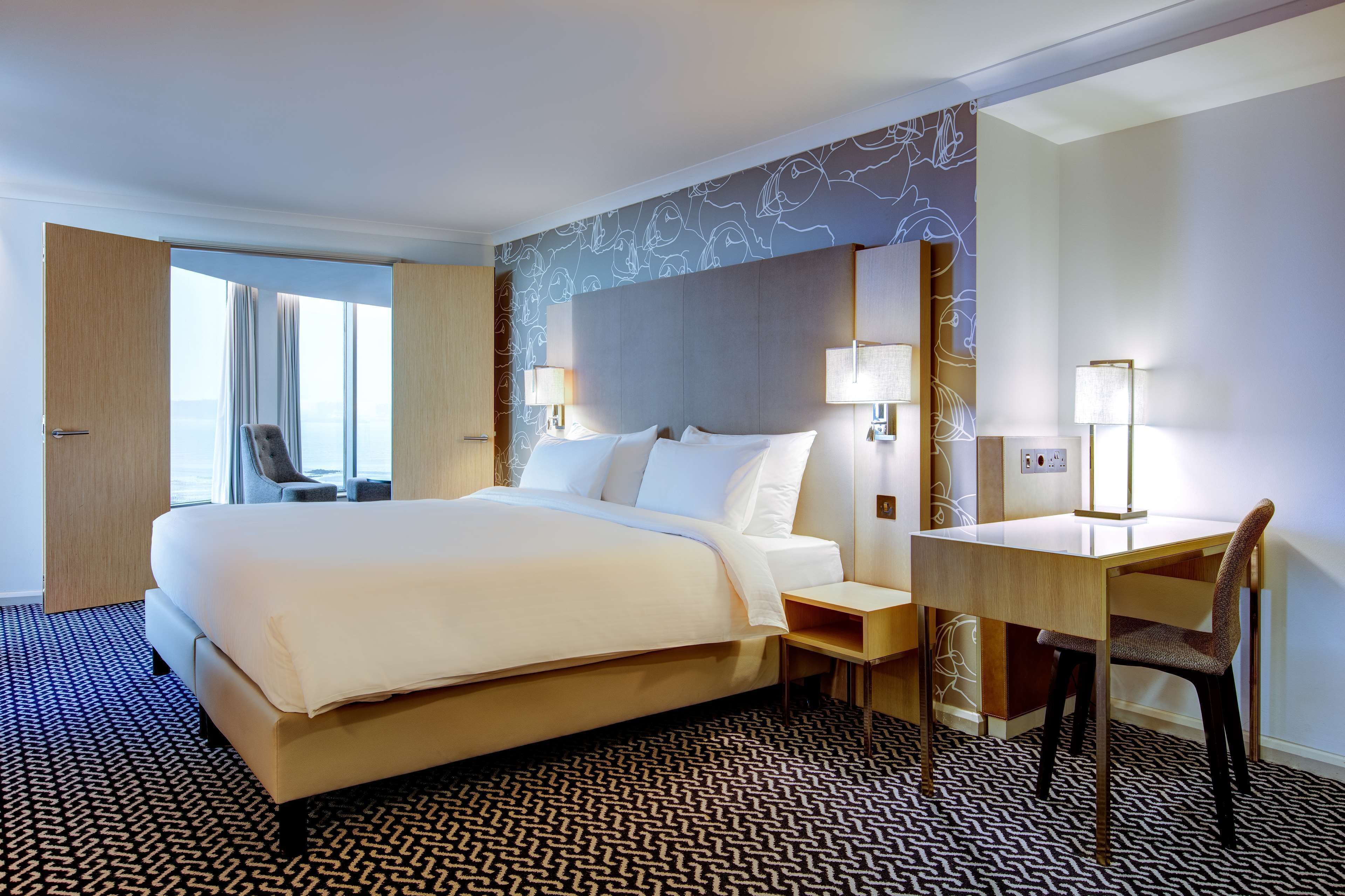 Habitación de hotel con una cama grande, un escritorio y una silla. Se ve una ventana con cortinas, una pared con diseño y una lámpara sobre el escritorio.