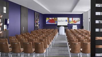 Uma sala de conferências com fileiras de cadeiras marrons, um palco com uma mesa branca e uma tela grande exibindo 'Radisson Blu'.