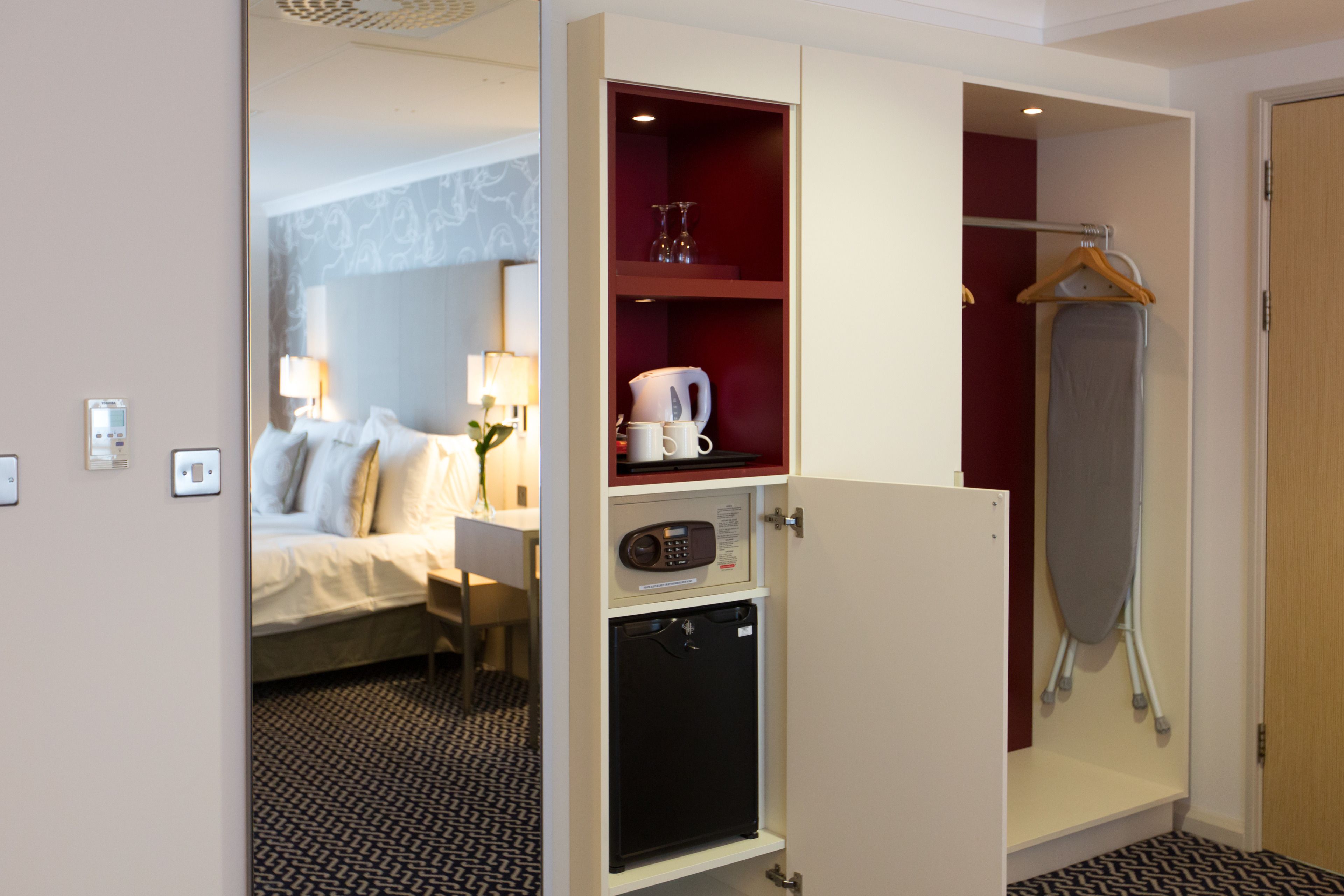 A hotel room with a bed, a desk with a lamp, a minibar, a shower area, and a wardrobe.