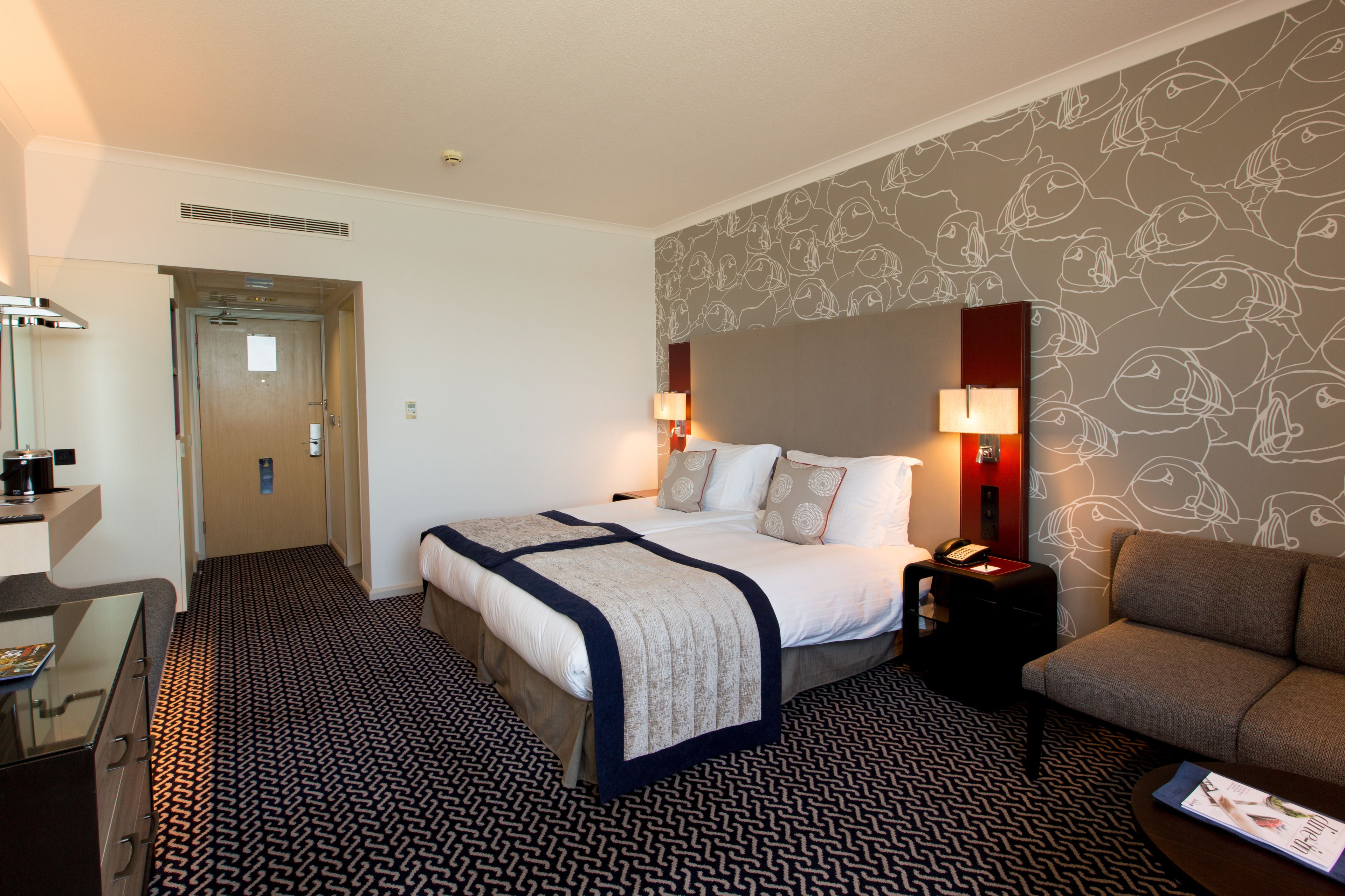 Una habitación de hotel con cama, mesita de noche, lámpara y un sofá.