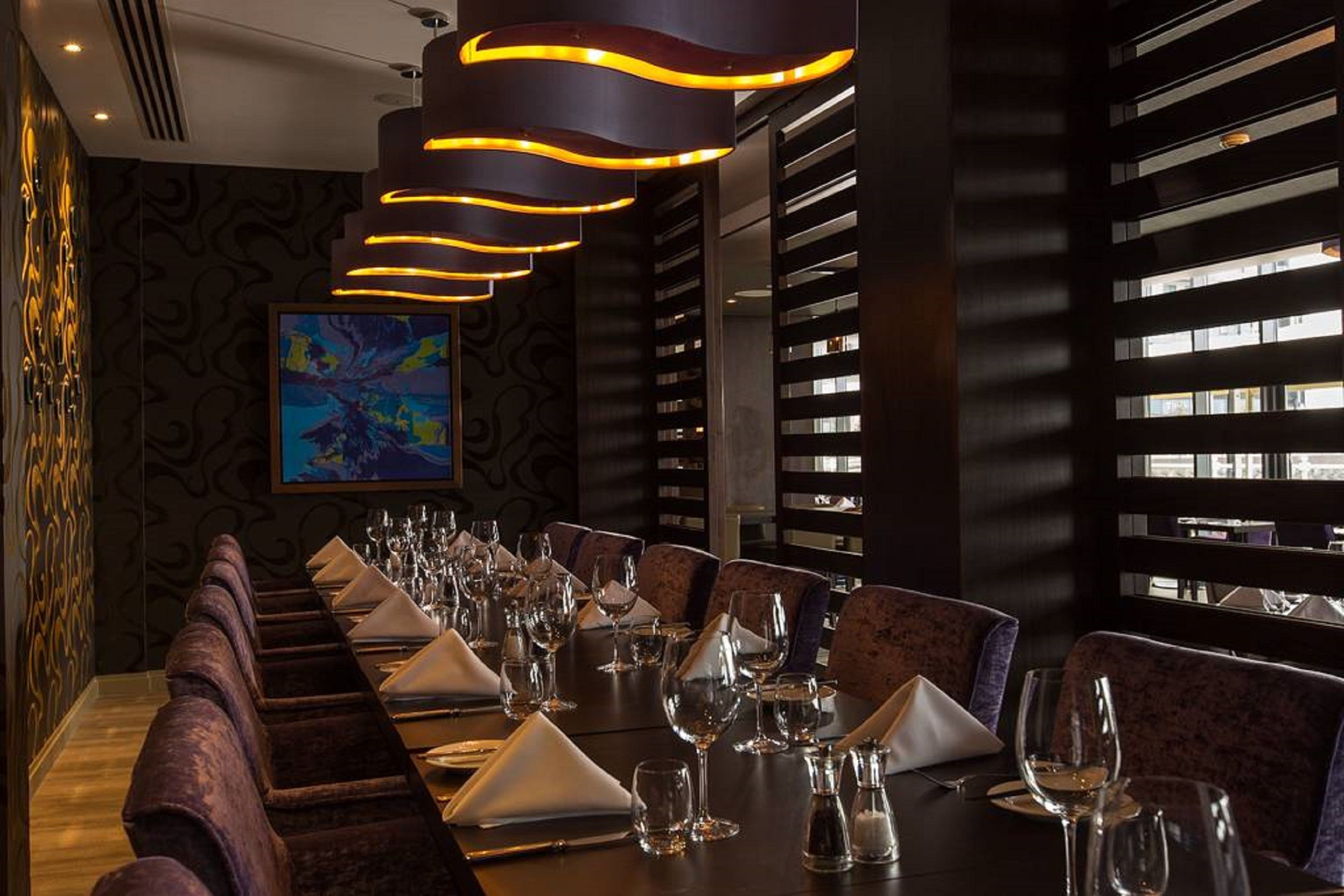 A long dining table set with multiple chairs, a modern chandelier, and a large abstract painting on the wall.