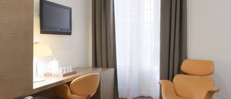 A hotel room with a wooden desk, two orange chairs, a television mounted on the wall, and a window with curtains.