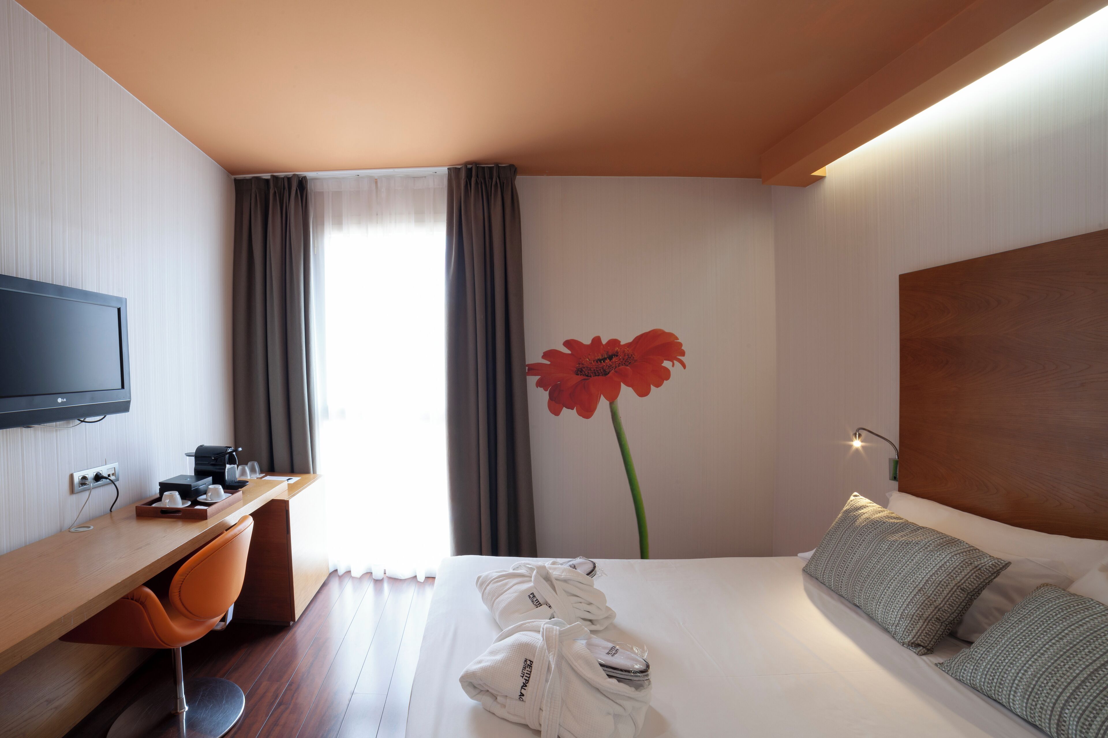 A hotel room with a wooden desk, a television, a bed with pillows, and a large red flower on the wall.