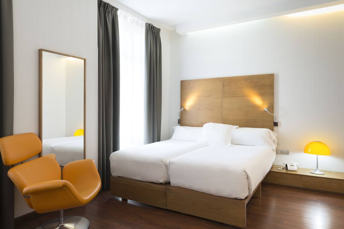 A hotel room with a double bed, a wooden headboard, a bedside table with a lamp, a chair, and a mirror.