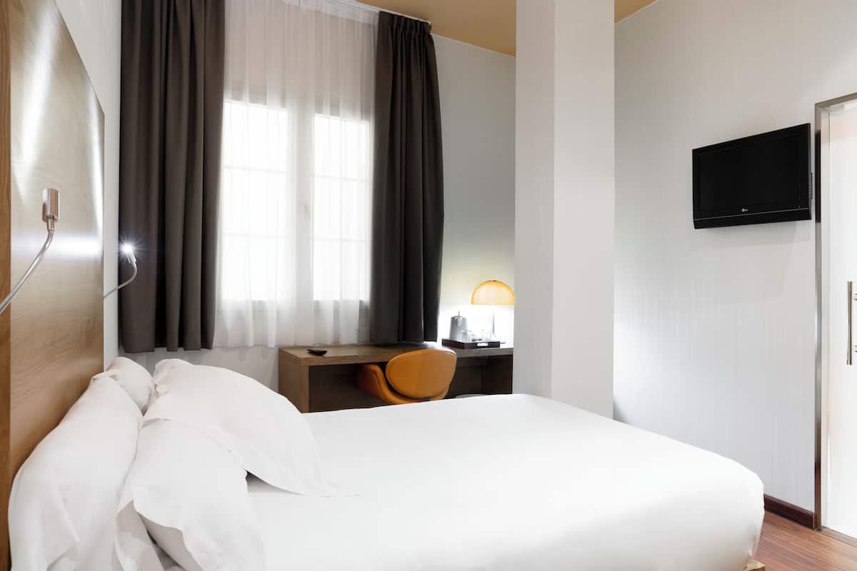 A hotel room with a bed, a desk with a chair, a TV, and a window with curtains.