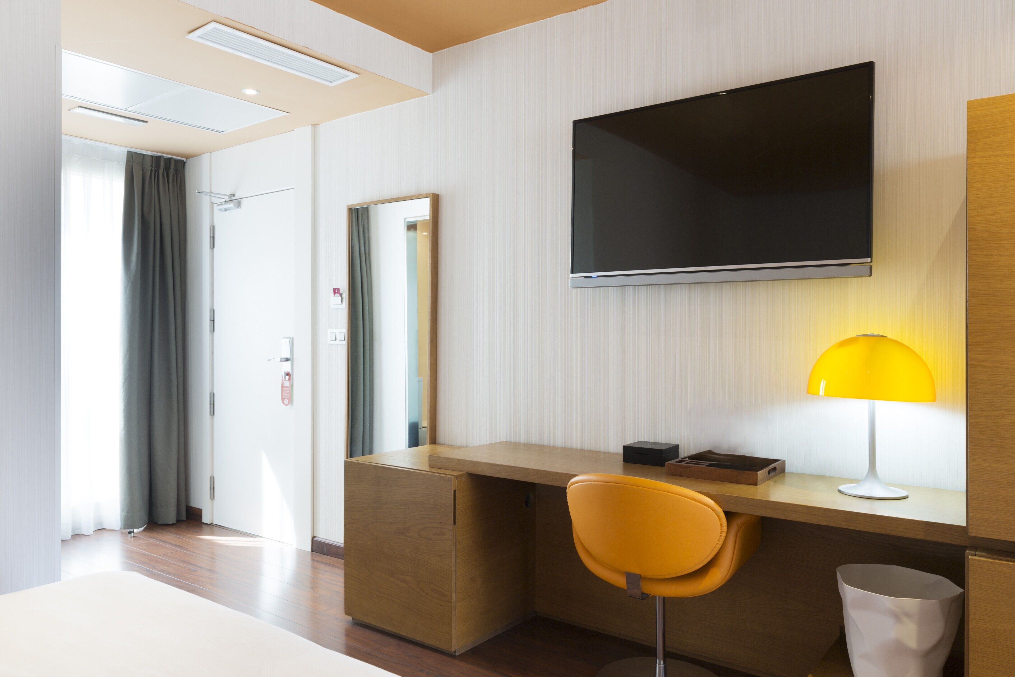 Une chambre d’hôtel moderne avec un bureau en bois, une chaise jaune, un téléviseur à écran plat fixé au mur et une lampe de chevet.