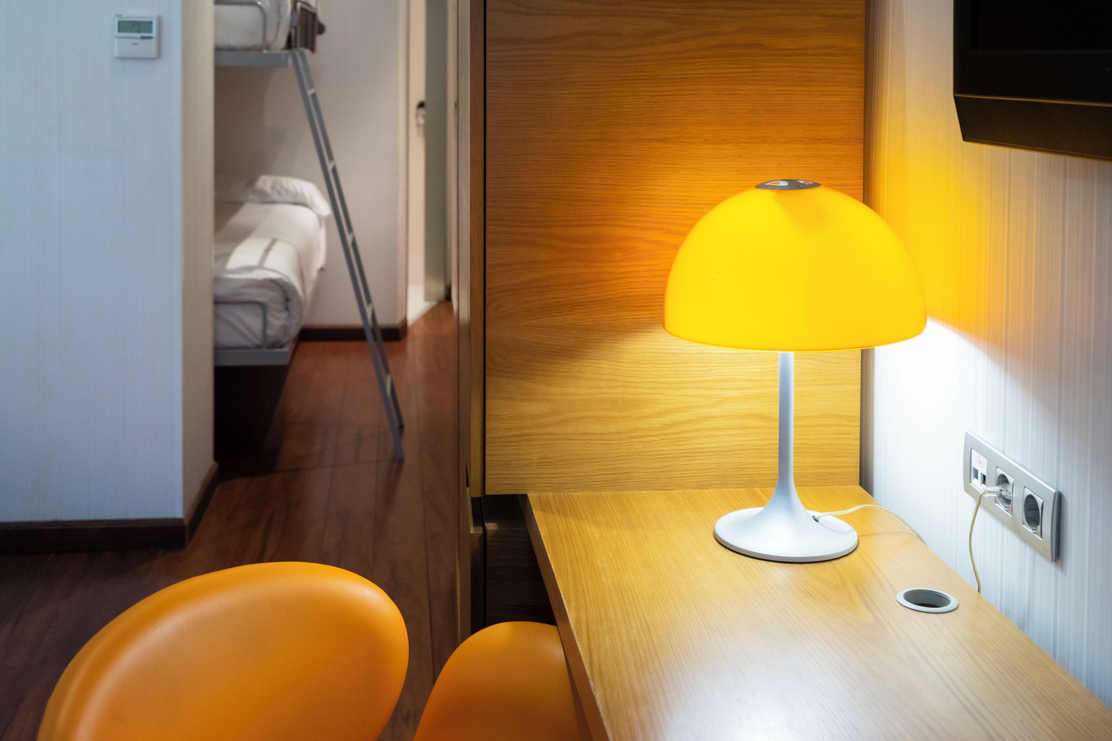 Une chambre d’hôtel avec un plancher en bois, un lit avec une literie blanche, une lampe jaune posée sur une table en bois et une chaise orange.