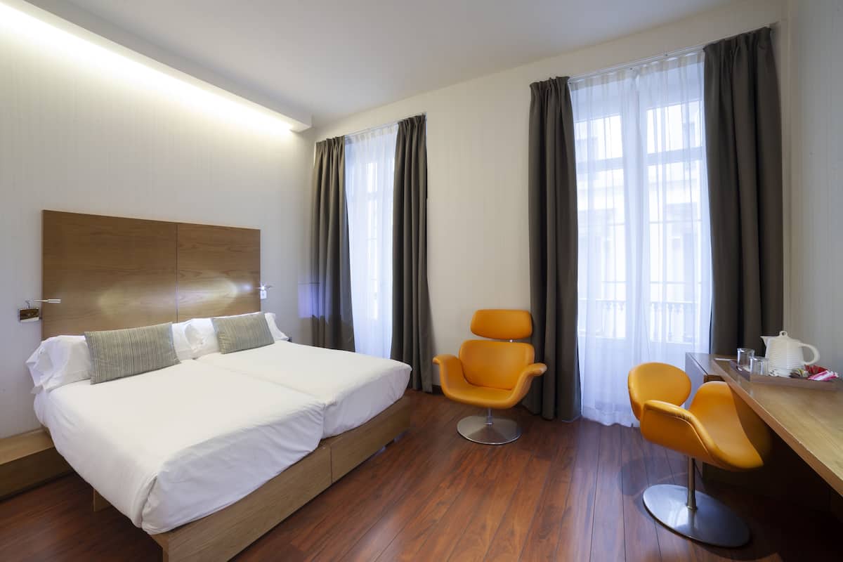 A hotel room with a large bed, two orange chairs, a wooden desk, and a window with curtains.