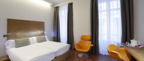 A hotel room with a large bed, two orange chairs, a wooden desk, and a window with curtains.