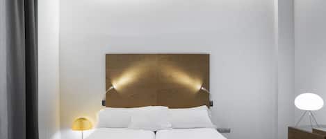 A modern hotel room with a wooden headboard, two beds, a bedside table with a lamp, and a nightstand with a lamp.