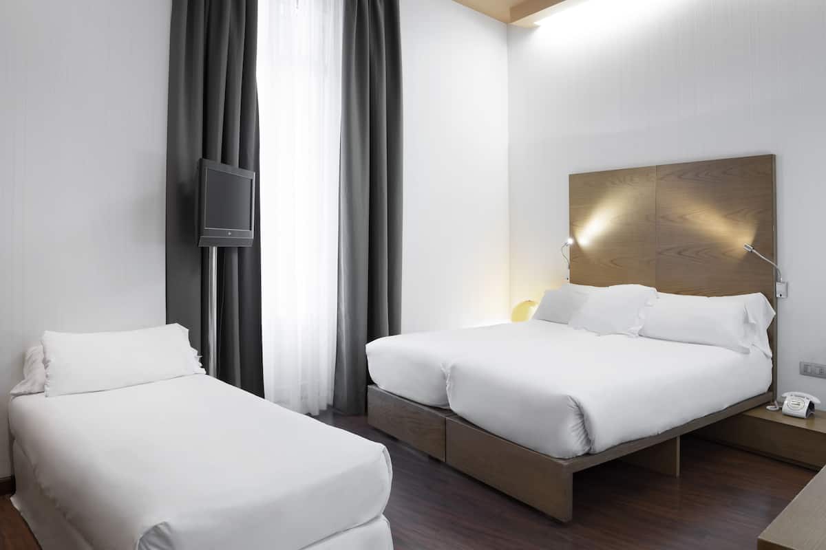 A hotel room with two beds, a TV mounted on the wall, a bedside table with a lamp, and a wooden headboard.