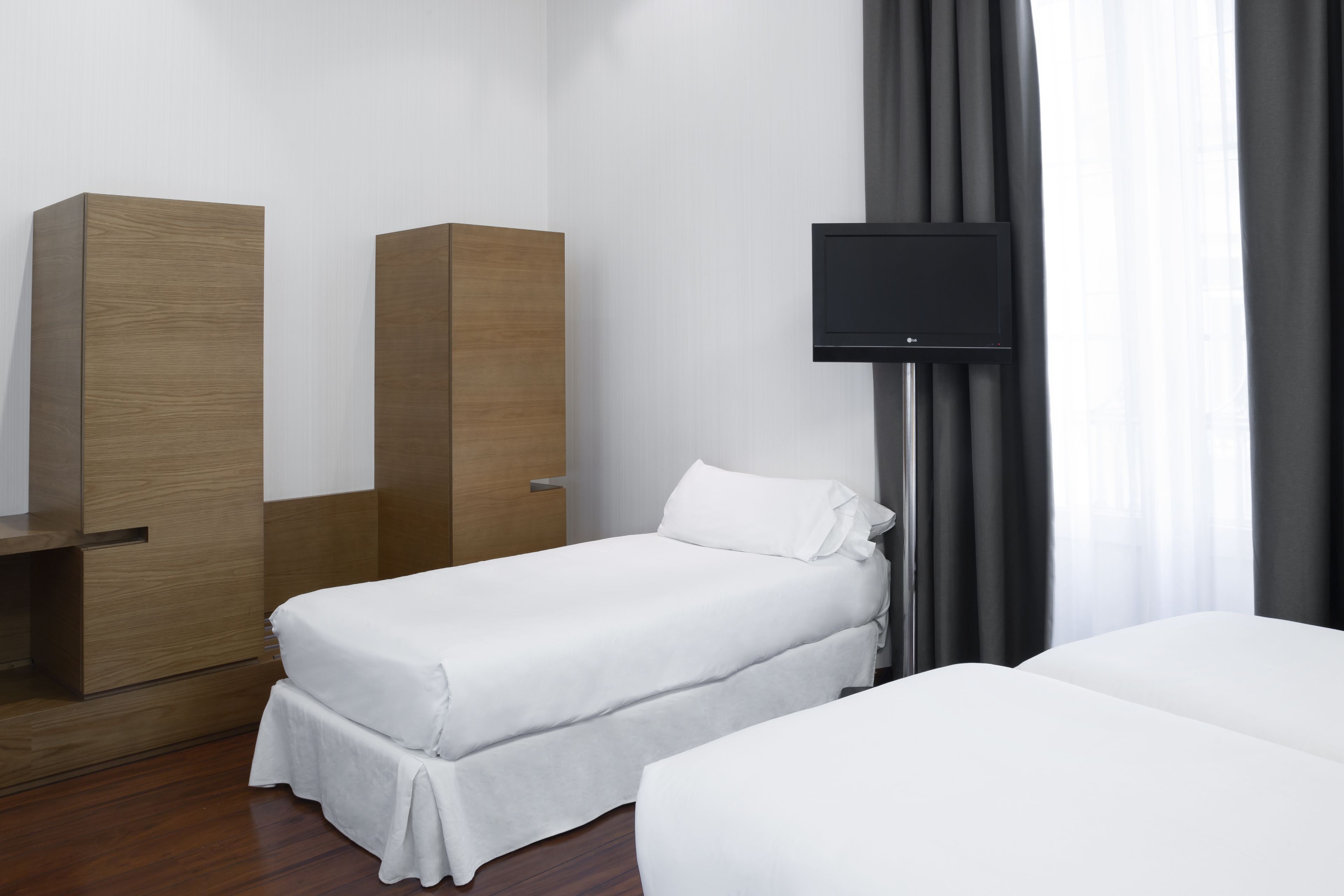 Habitación de hotel con una cama individual, un armario de madera, un televisor sobre un mueble bajo y una ventana con cortinas.
