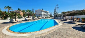 A swimming pool with lounge chairs and umbrellas, surrounded by palm trees and a building with a white facade. - Horizon Resort (Santorini)