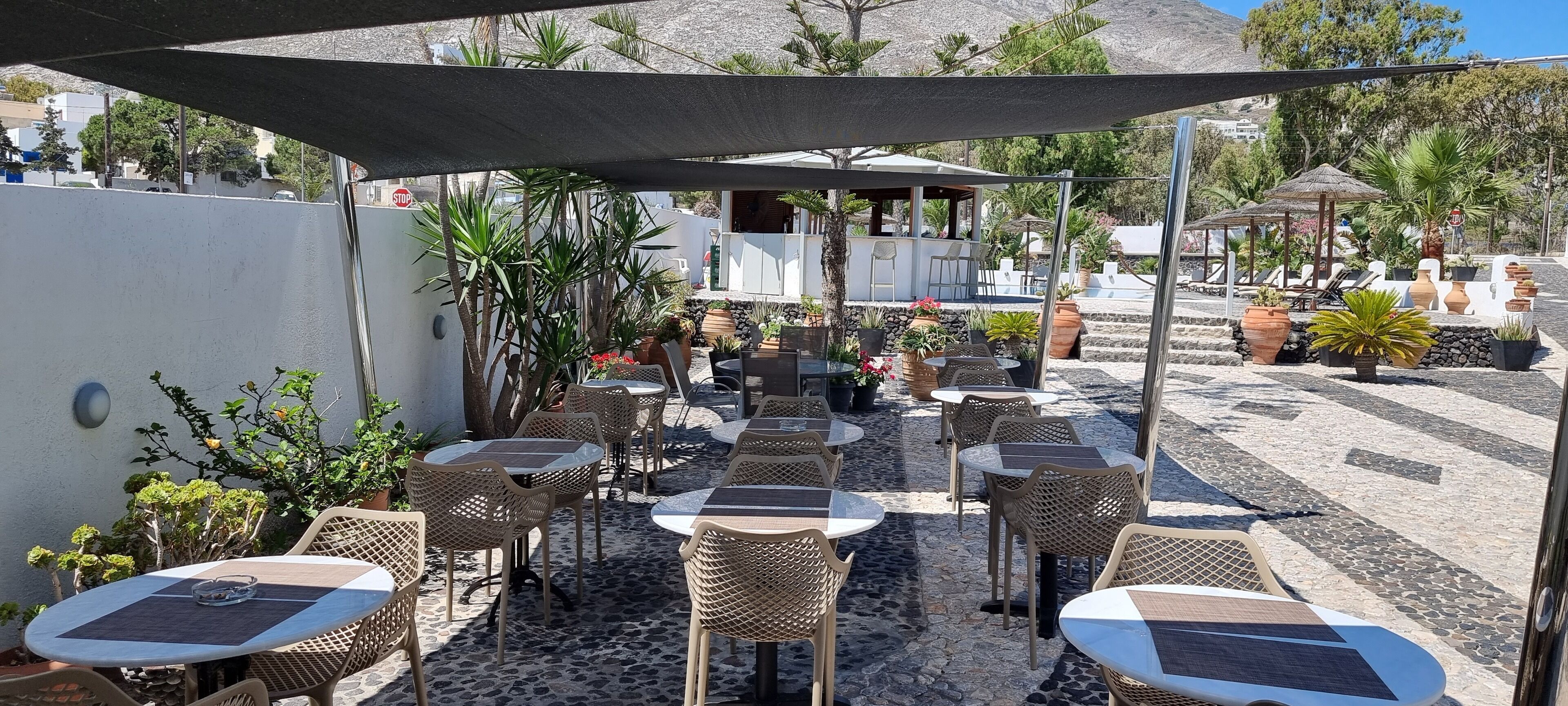 Une terrasse extérieure avec des tables rondes et des chaises en osier, entourée de plantes en pot et d’un gazebo à toit de chaume.