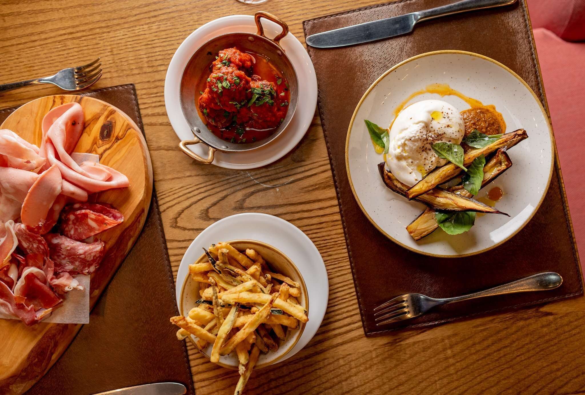 Una mesa de madera con una variedad de alimentos, incluyendo embutidos, un bol de papas fritas, un plato de albóndigas en salsa de tomate, y una fuente con un huevo pochado y verduras.