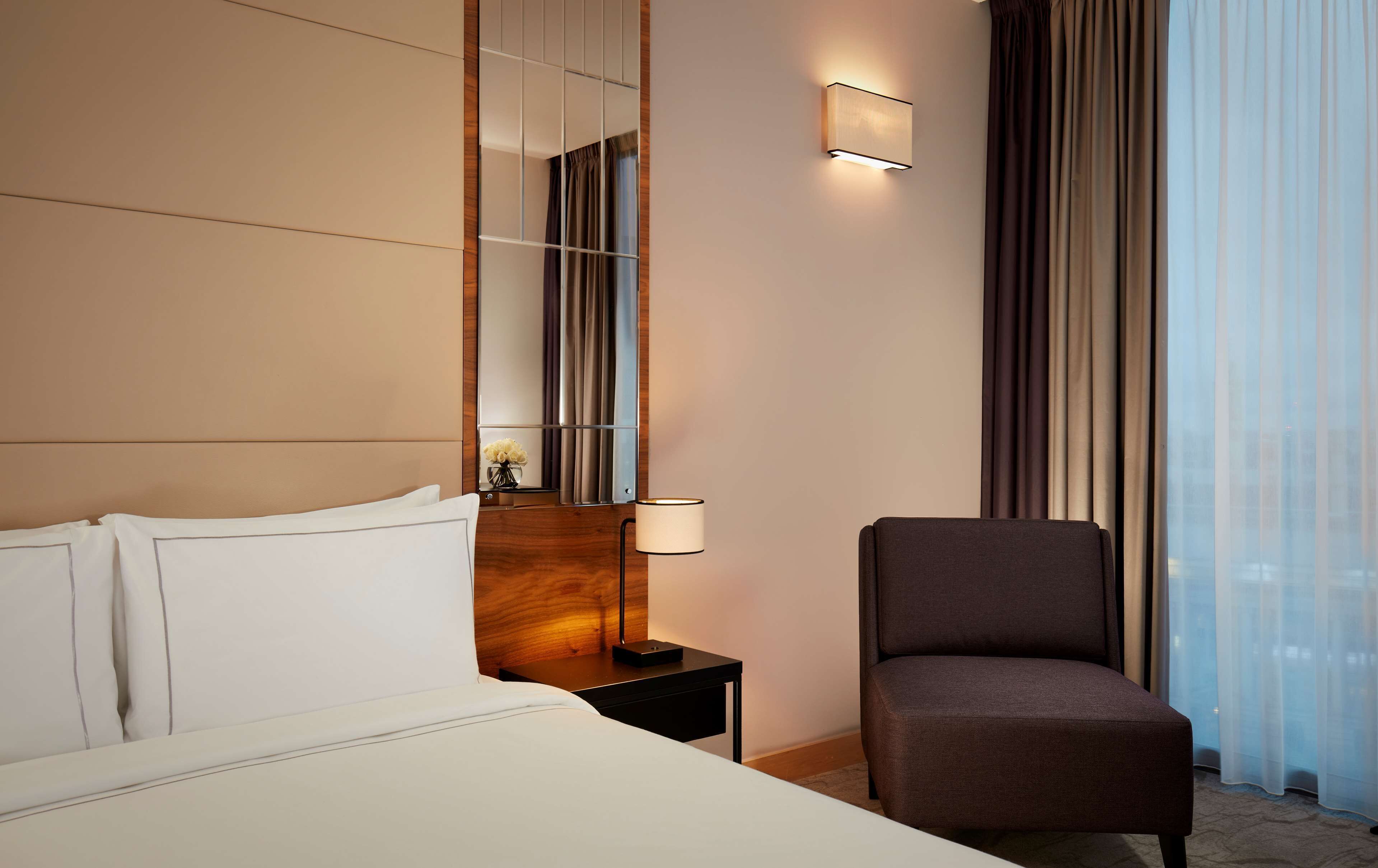 Una habitación de hotel con una cama, una silla, una mesita de noche, una lámpara, un espejo y una ventana con cortinas.