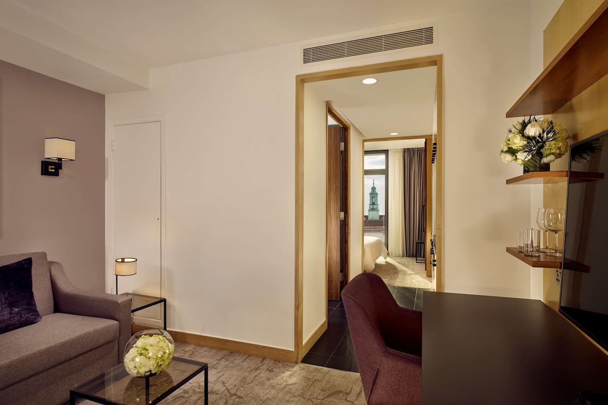 A modern hotel room with a sofa, a coffee table with a vase, a lamp, a mirror, and a wooden shelf with glasses.