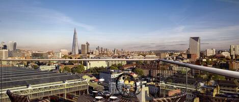 Une vue panoramique d’une ville, avec un gratte-ciel imposant qui se détache à l’horizon, un pont et une terrasse sur le toit aménagée avec des meubles en osier.