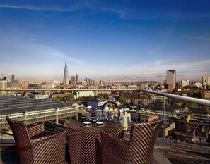 Une vue panoramique d’une ville, avec un gratte-ciel imposant qui se détache à l’horizon, un pont et une terrasse sur le toit aménagée avec des meubles en osier.