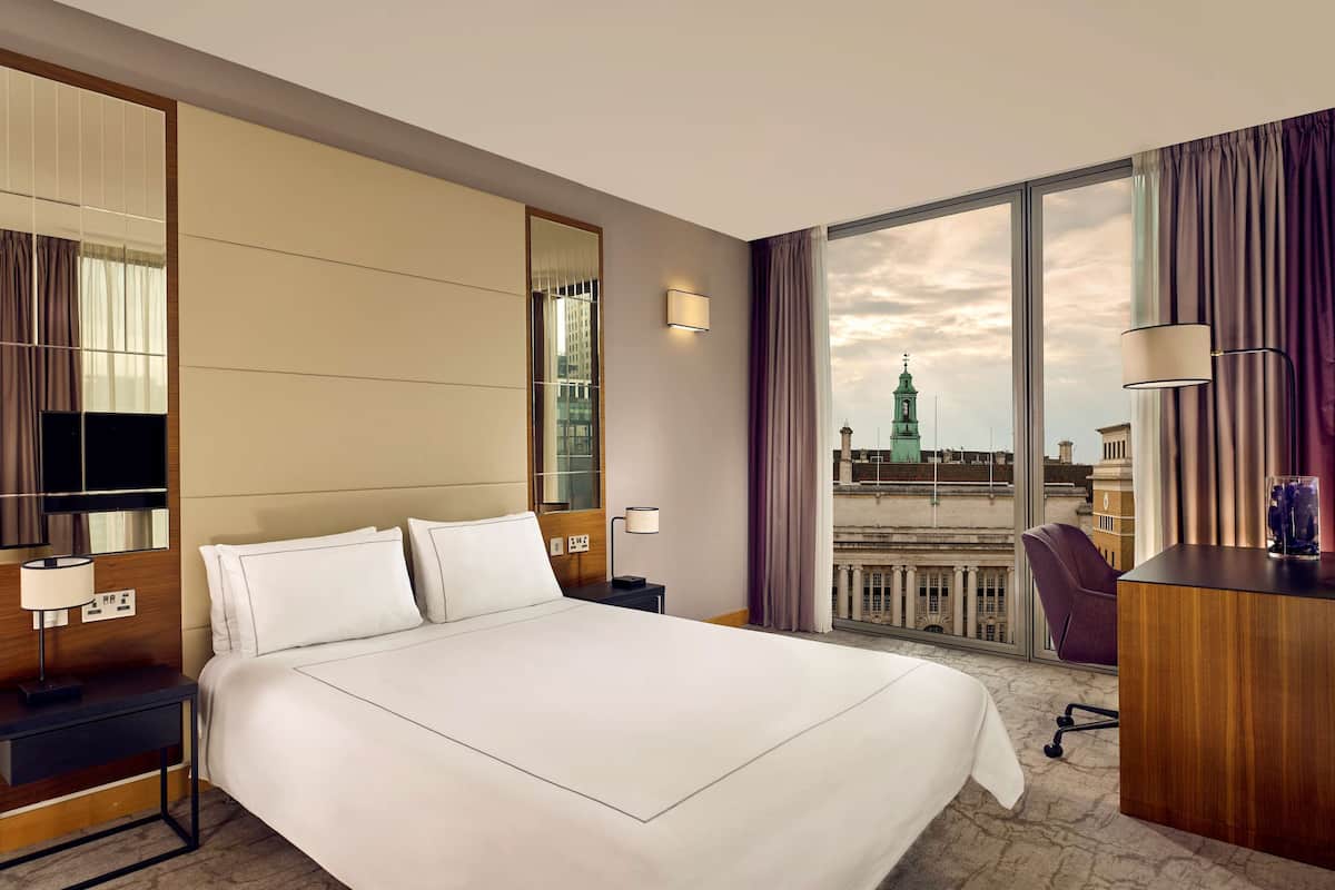 A hotel room with a large bed, a desk with a lamp, a chair, a TV, and a large window with a city view.