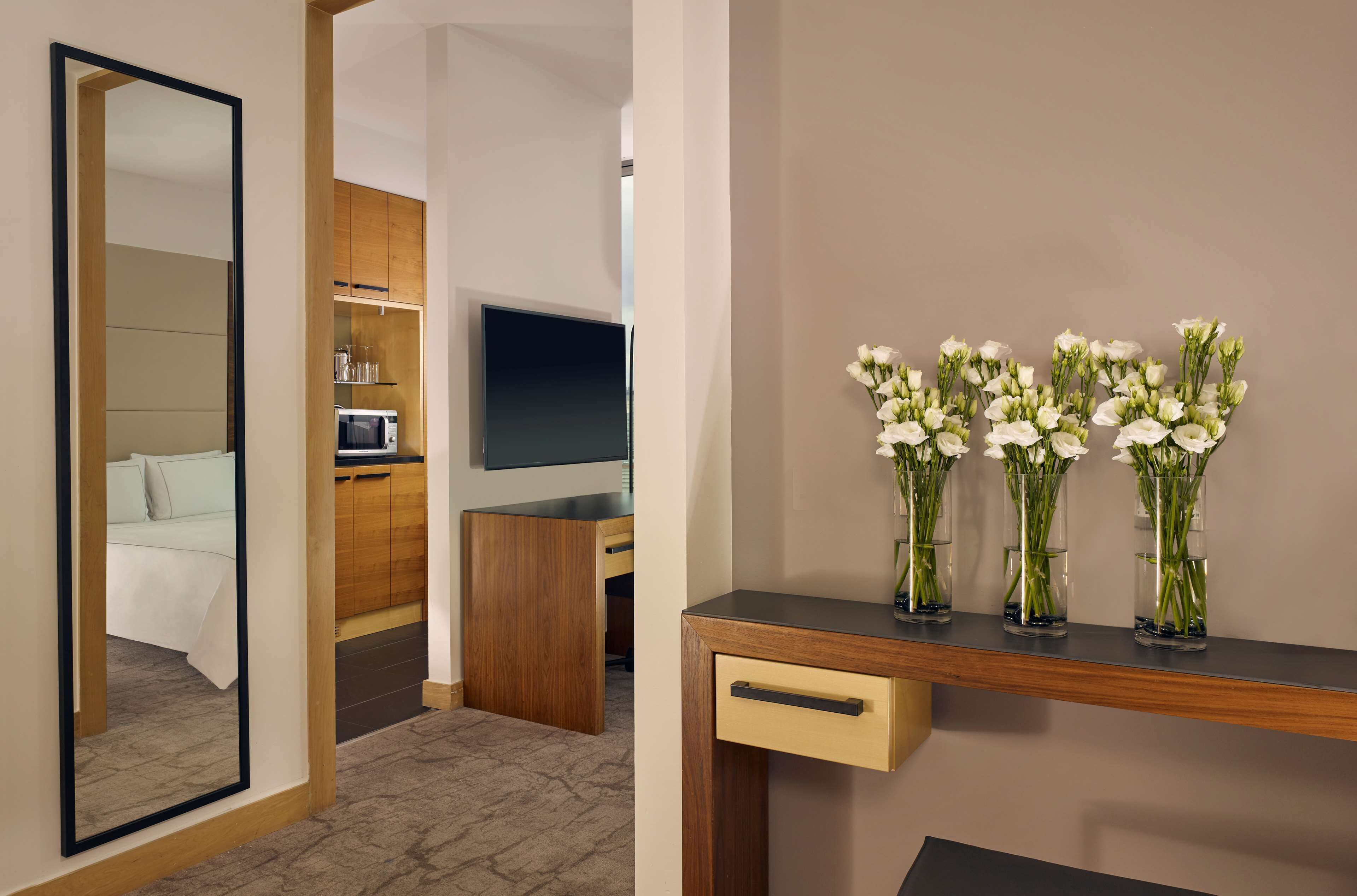 A hotel room with a bed, a wooden desk, a television, and a wall-mounted shelf with vases of flowers.
