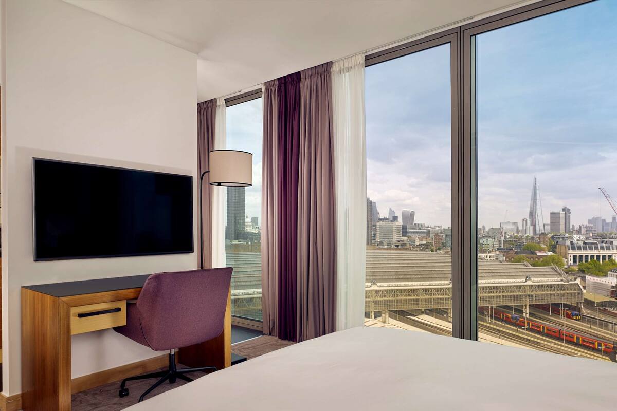 A hotel room with a large window, a flat-screen TV, a desk, a chair, and a bed.