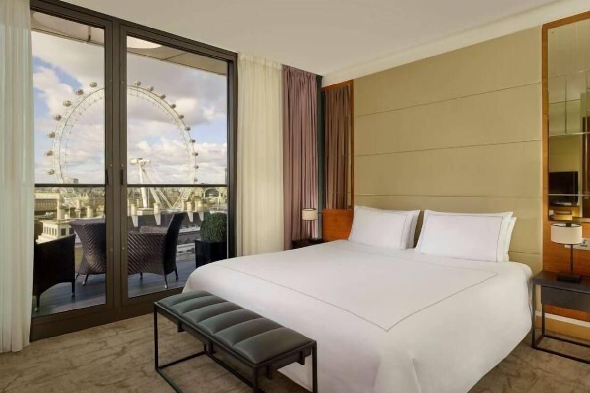 A hotel room with a large bed, a bench, a view of a Ferris wheel, and a balcony with chairs.