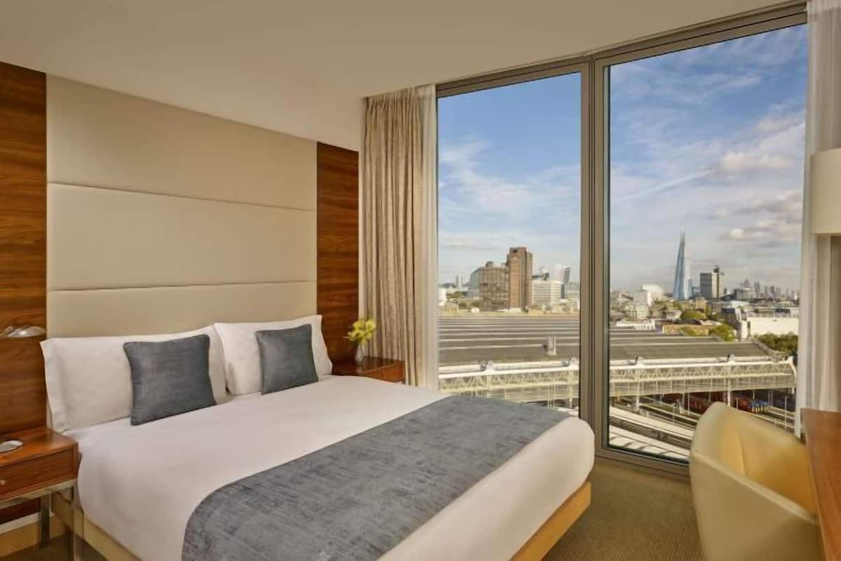 A hotel room with a large bed, a nightstand, a chair, and a view of a cityscape through large windows.