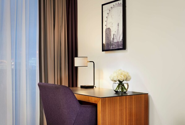 A hotel room with a wooden desk, a purple chair, a lamp, and a framed picture of a Ferris wheel. - Park Plaza County Hall London (London)