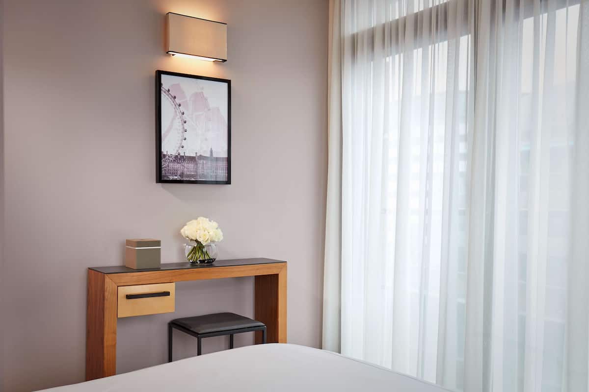 A hotel room with a bed, a wooden console table with a vase of flowers, a framed picture on the wall, and a window with sheer curtains.