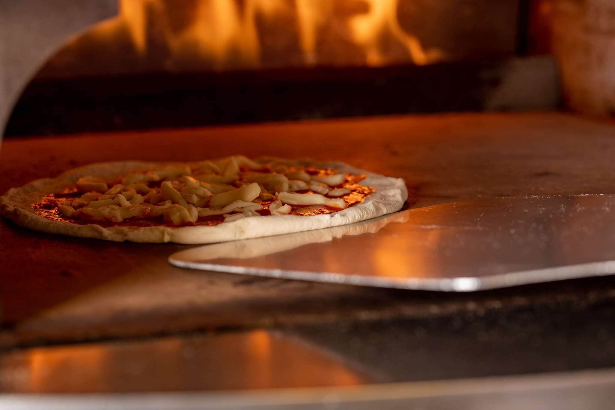 A pizza on a metal tray in front of a wood-fired oven.