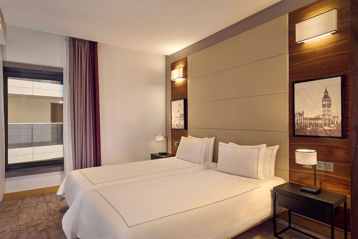 A hotel room with a large bed, a desk, a chair, and a view of a cityscape through a window.