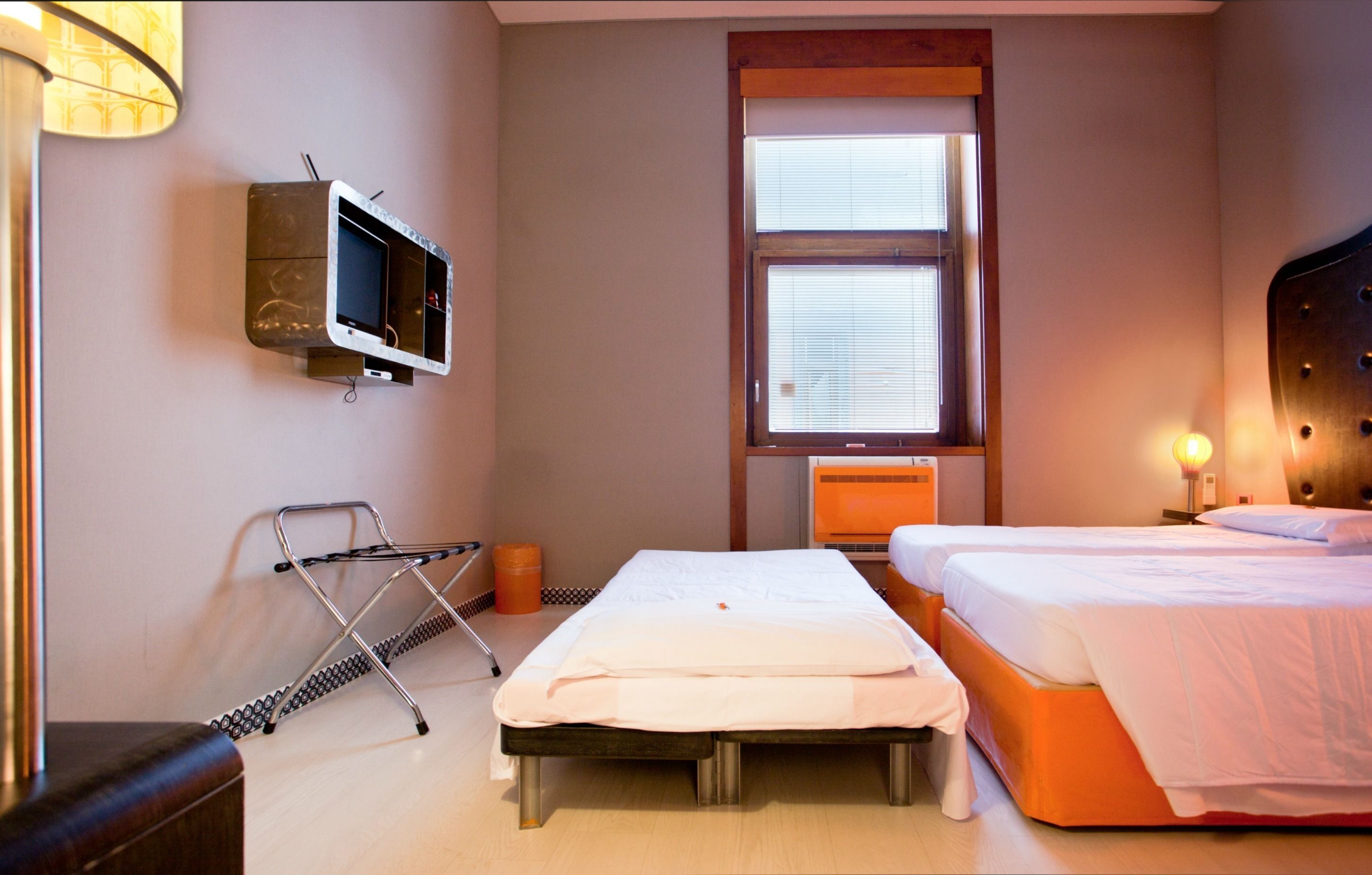 A hotel room with two beds, a television mounted on the wall, a window with blinds, and a bedside table with a lamp.