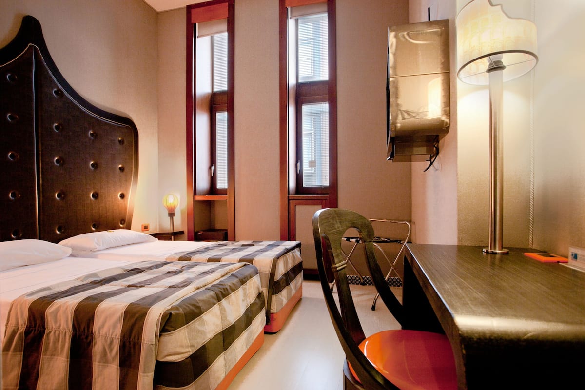 Een hotelkamer met twee bedden, een bureau, een stoel, een lamp en een raam met jaloezieën.