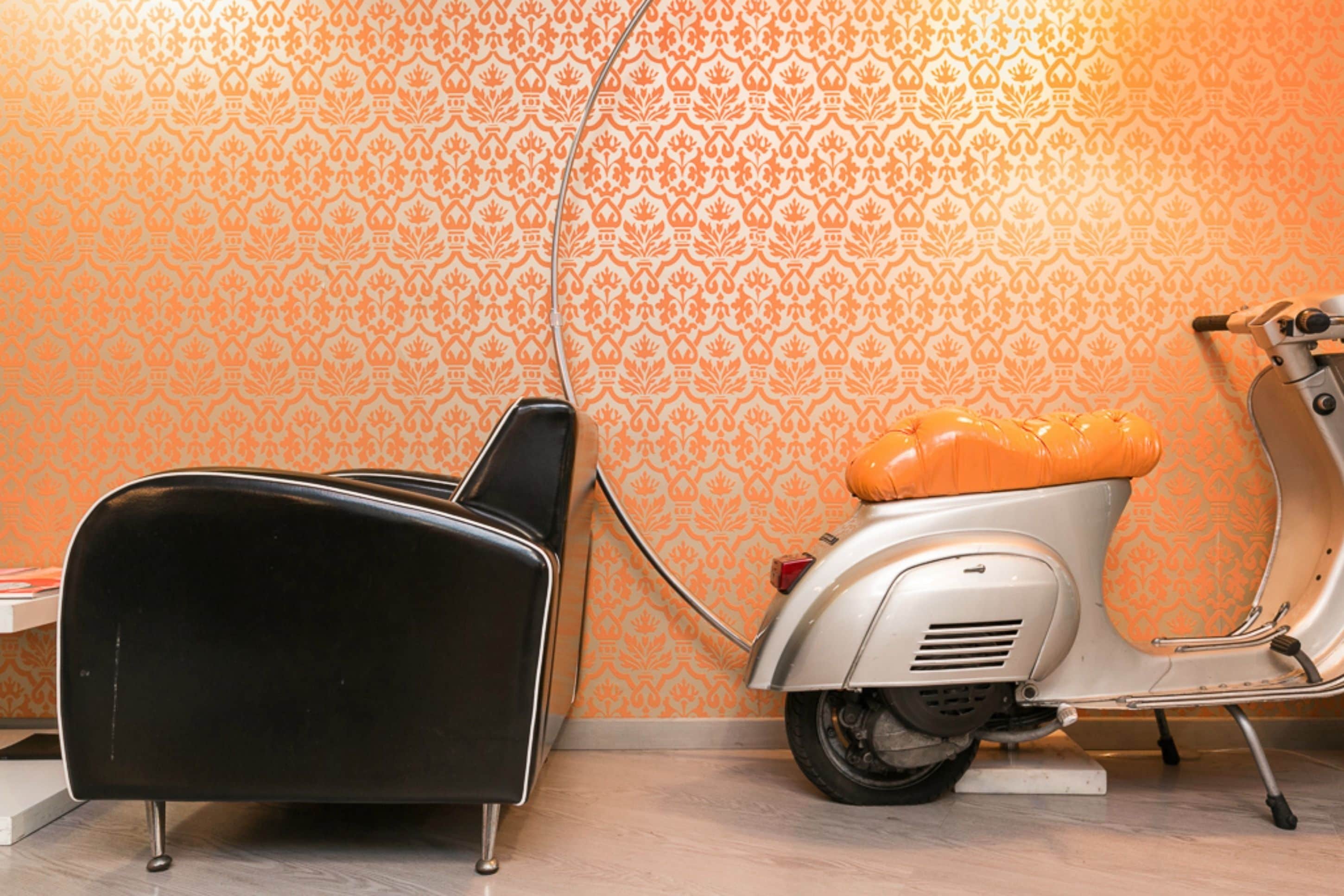 A room with a patterned orange wallpaper, a black chair, and a white and orange vintage scooter.