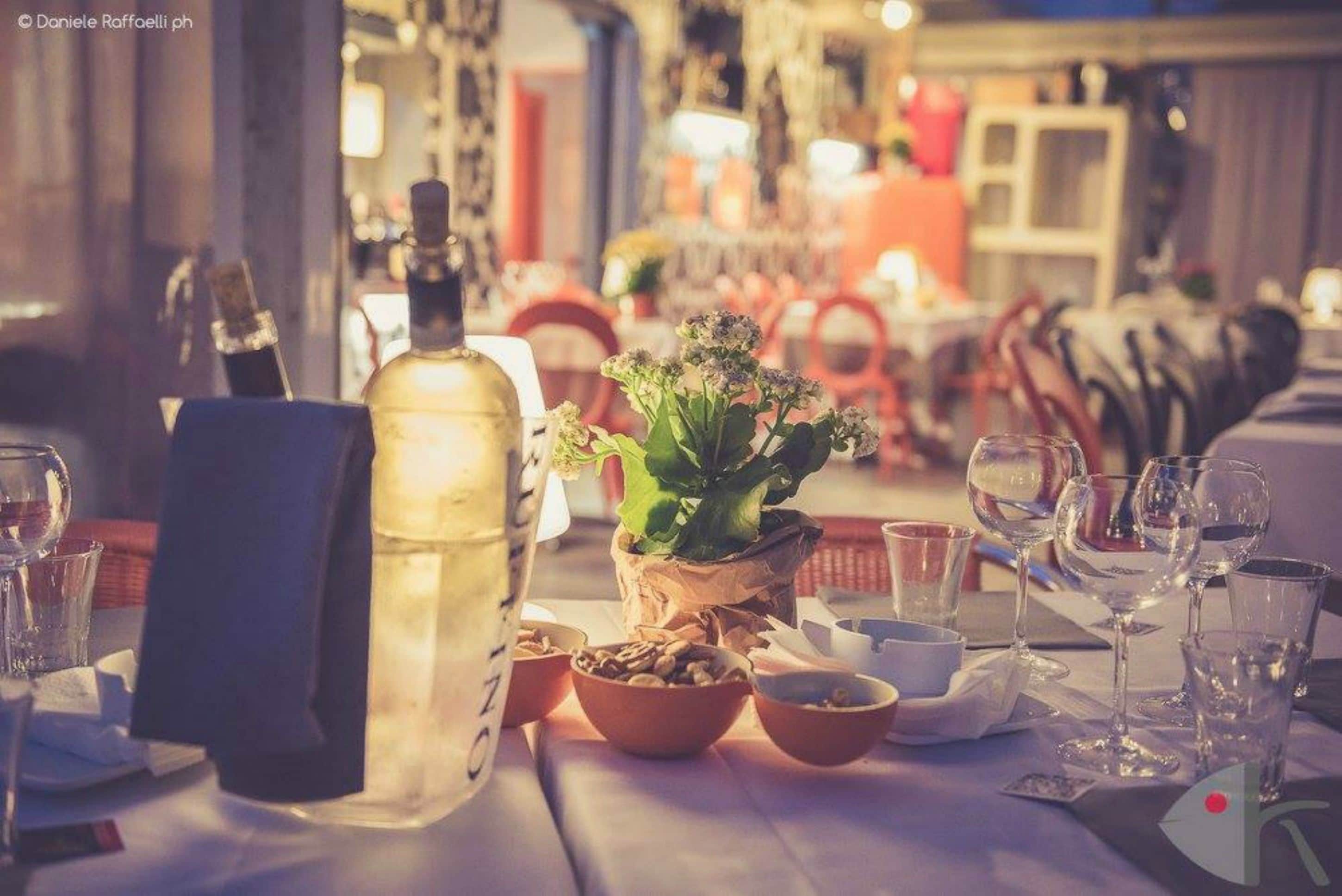 Ein Esstisch ist gedeckt mit einer Flasche, Schüsseln und Weingläsern, im Hintergrund steht ein Stuhl und ein Blumentopf.
