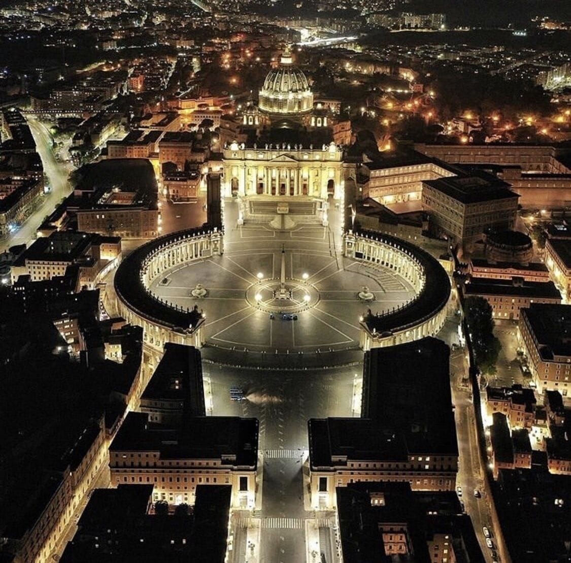 Nachtansicht von oben eines großen, kreisförmigen Platzes mit einem zentralen Brunnen, umgeben von klassischer Architektur und beleuchteten Gebäuden.