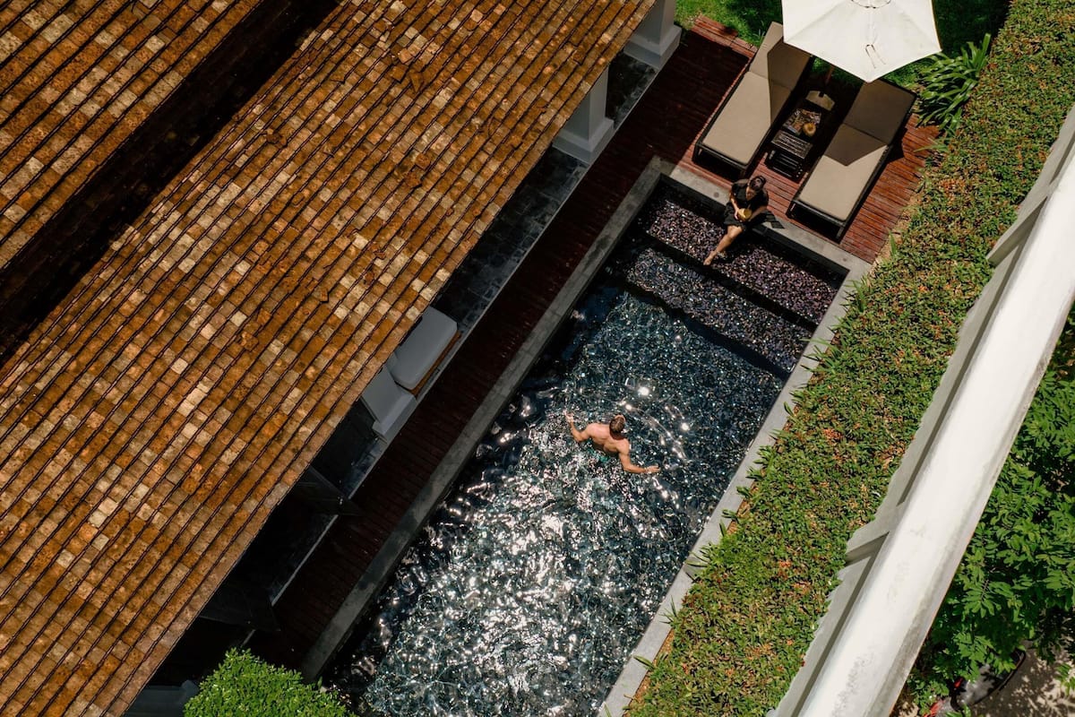 Ein Blick von oben auf ein Schwimmbecken mit einer Person, einer Liegewiese mit Liege und Sonnenschirm sowie einem Backsteinbau.
