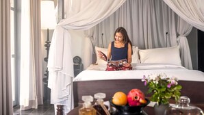 A room with a bed, curtains, a bedside table with a lamp, and a window with curtains. - Sofitel Luang Prabang (Luang Prabang)
